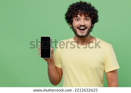 Young excited happy Indian man 20s in basic yellow t-shirt hold in hand use mobile cell phone with blank screen workspace area isolated on plain pastel light green background People lifestyle concept