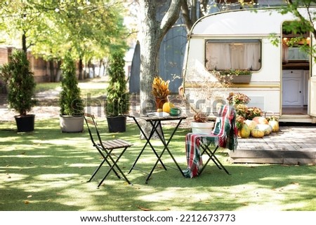 Autumn garden with chair and table. Backyard with orange pumpkin. Interior cozy campsite with flowers and pumpkins. Wooden RV house porch with garden furniture. Campsite with caravan trailer in garden