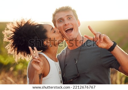Funny, kiss and couple bonding during vacation on a farm with peace sign. Happy interracial husband and wife standing close together on vineyard in summer. Young husband and wife feeling in love