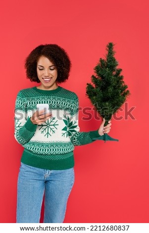 smiling african american woman in warm sweater holding small christmas tree and mobile phone isolated on red