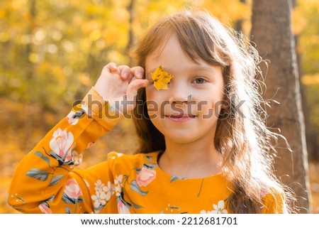 Close portrait of a little girl. She covers her eye with a maple autumn leaf. Fall season and children concept.