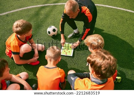 Football training. Soccer coach explaining game rules and strategy using tablet, map. Sports junior team sitting on grass pitch with trainer. Concept of sport, achievements, studying, goals, skills Royalty-Free Stock Photo #2212683519