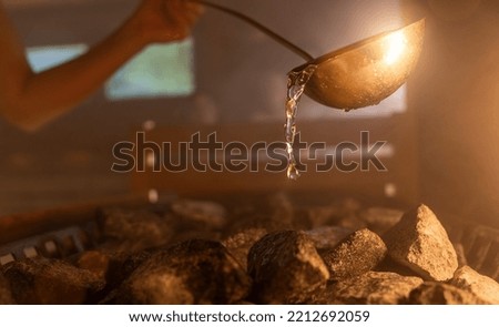 Woman pouring water onto hot stone in sauna room with a group of people. Steam an water on the stones, spa and wellness concept, relax in hot finnish sauna. Royalty-Free Stock Photo #2212692059