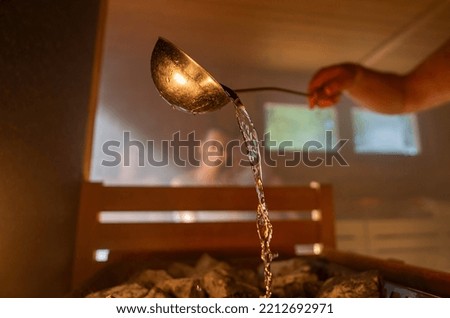 Man pouring water onto hot stone with metal spo in sauna room with a group of people. Steam an water on the stones, spa and wellness concept, relax in hot finnish sauna. Warm temperature bath therapy. Royalty-Free Stock Photo #2212692971