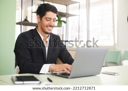 Optimistic ethnic man in formal business wear using laptop sitting on the workplace in modern open space office, young hispanic male employee replying emails, working on a new project Royalty-Free Stock Photo #2212712871