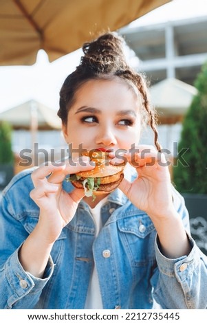 A young glamorous woman with dreadlocks and red lipstick is sitting and eating a burger in a street cafe, the concept of eating. long-lasting lipstick