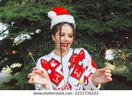 a woman in a winter sweater, with a Santa Claus hat and red lips holds a sparkler against the background of Christmas trees. new year christmas