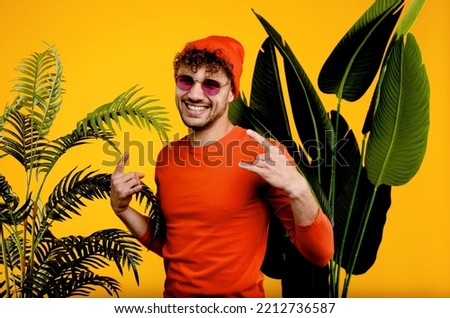 Portrait of young attractive funky man showing rock metal sign, isolated yellow background with green plants . High quality photo
