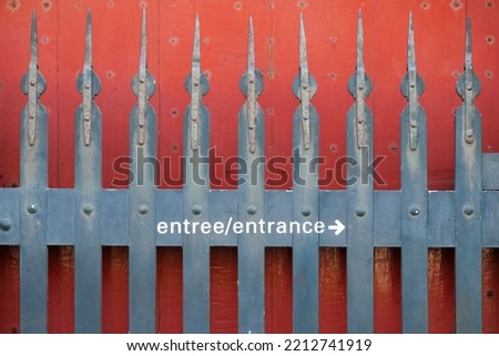 Gate, fence with spike and letters saying 'entree, entrance' with an arrow pointing to the right. Entrance to the right. No entrance here. Closed gate with red background. Closed, spiked gate.  