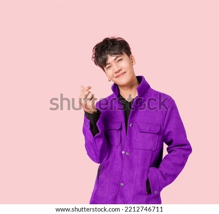 Portrait of a happy Asian handsome man in fashionable purple jacket clothing standing smiling using fingers to make a mini heart sign isolated on pink background. young male fashion model lifestyle.
