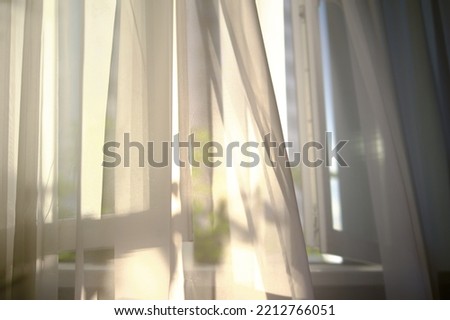 wind blows through the open window in the room. white curtain veil from an open window. Sunny day, the sun's rays sunlight penetrate see transparent tulle the room. fresh air fills the room. Royalty-Free Stock Photo #2212766051