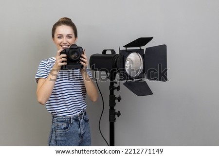 Portrait of smiling satisfied woman photographer wearing striped T-shirt standing with photocamera and reflector, looking at camera. Indoor studio shot isolated on gray background.