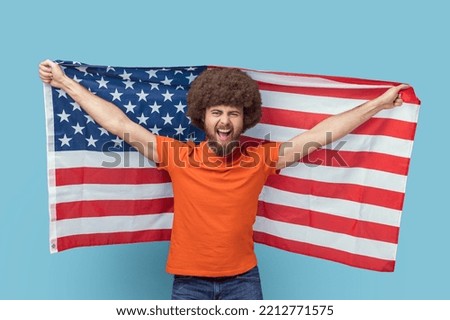Portrait of man with Afro hairstyle in T-shirt holding USA flag and looking at camera with rejoicing look, screaming, celebrating national holiday. Indoor studio shot isolated on blue background.