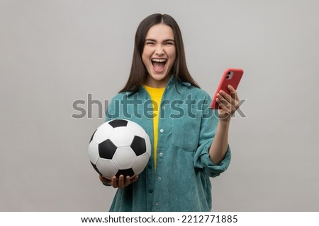 Extremely happy excited woman standing holding soccer ball and using smart phone, betting and winning, wearing casual style jacket. Indoor studio shot isolated on gray background.