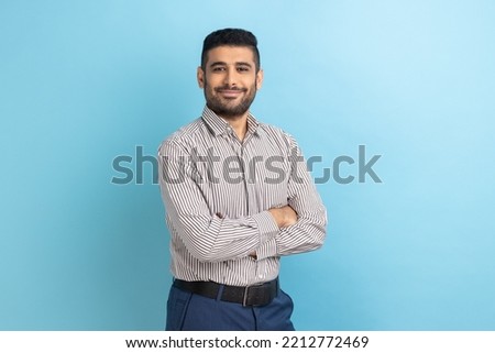 Young happy successful businessman posing with happy confident expression, looking at camera, keeping arms folded, wearing striped shirt. Indoor studio shot isolated on blue background.