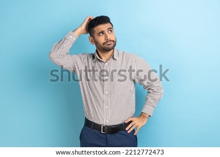 Portrait of young pensive businessman with beard touching head and thinking about important question, making hard decision, wearing striped shirt. Indoor studio shot isolated on blue background. Royalty-Free Stock Photo #2212772473