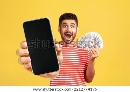 Surprised happy man with beard in red T-shirt, looking with big amazed eyes and open mouth, holding money and showing mobile phone with empty screen. Indoor studio shot isolated on yellow background. Royalty-Free Stock Photo #2212774195