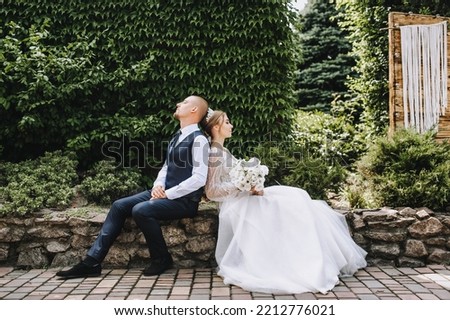 A stylish, young groom and a beautiful bride in a white dress with a bouquet in their hands, a diadem on their heads, sit in a park in nature. Wedding photo of lovely newlyweds.
