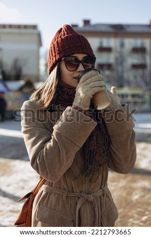 Woman in warm winter clothes and sunglasses drinking hot coffee from takeaway paper cup while enjoying sunny day in snowy city 