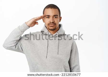 Image of young man saluting, holding hand near forehead, army greeting, standing over white background