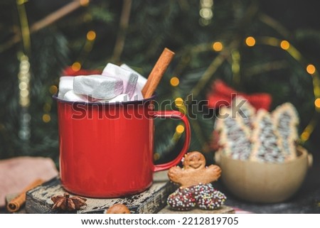 One red iron mug with hot cocoa, marshmallows on top with cinnamon, gingerbread man on cutting board on blurred background with garland, side view close-up. Holiday drink concept.
