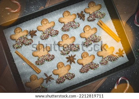 Three rows of gingerbread men with chocolate and sprinkles on white baking paper in a baking dish with cinnamon, candy cane, fir branches and golden bokeh on a dark cement background, top view close-