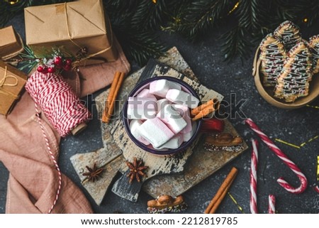 One red iron mug with hot cocoa, marshmallows on top with cinnamon, gingerbread man on a cutting board and christmas candy with decoration on a dark cement background with fir branches garland, top vi