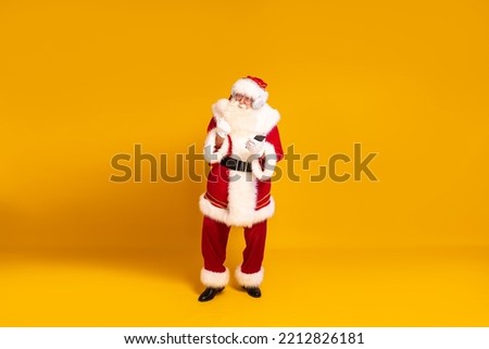 Happy bearded real Santa Claus with headphones dancing and listening music from mobile phone. Yellow studio background. Christmas time. Copy space. Xmas vibes. 
