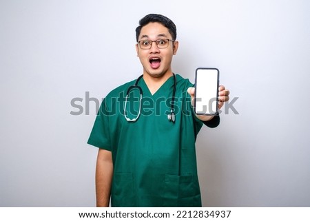 Asian young man nurse, doctor wearing scrubs and stethoscope looking to camera showing blank smartphone screen recommending application, isolated on white background