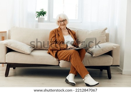 a lovely, joyful elderly woman, a businesswoman, is sitting on a cozy sofa near the window in a brown pantsuit and happily working on a laptop from home. Full-length photo