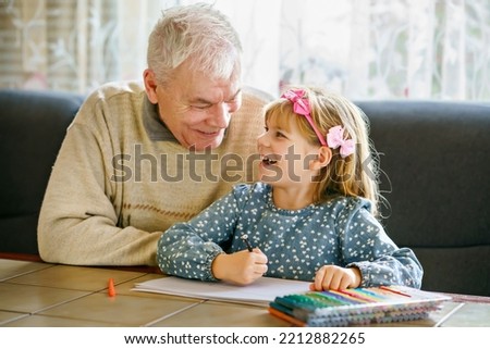 Cute little preschool girl and handsome senior grandfather painting with colorful pencils at home. Grandchild and man having fun together