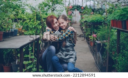 Young attractive worker of greenhouse and her cute emotional child are taking selfie in workplace hugging kissing and posing looking at smart phone camera.