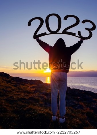 Silhouette of a teenager girl holding sign 2023 above her head. Sunrise sun in the background. Welcoming new year concept. Warm and cool tone.