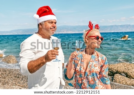 Funny adult couple wearing christmas costumes drinking sparkling wine and enjoying christmas vacations on tropical beach. Winter holiday concept.