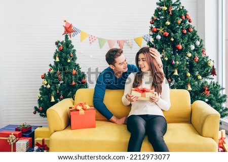 Young Asian man surprises her girlfriend with a Christmas gift at home with a Christmas tree in the background. Image with copy space.