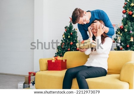 Young Asian man surprises and closes her eyes with a Christmas gift at home with a Christmas tree in the background. Image with copy space.