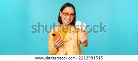 Online shopping. Stylish young asian woman in sunglasses, showing credit card and using smartphone, paying in internet, making purchase, standing over blue background