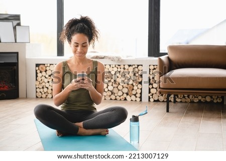 Happy relaxing african woman young athlete resting on fitness mat in lotus yoga position using smart phone for sport tracking application online after training at home Royalty-Free Stock Photo #2213007129