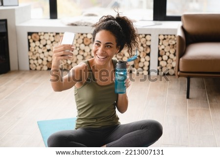 Home workout. Online vlogging blogging training on fitness mat using cellphone. African young female athlete doing yoga exercises at home, having videocall, video tutorial, taking selfie Royalty-Free Stock Photo #2213007131