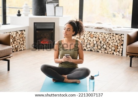 Pensive thoughtful relaxing african woman young athlete resting on fitness mat in lotus yoga position using smart phone for sport tracking application online after training at home Royalty-Free Stock Photo #2213007137