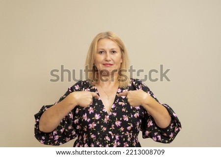 Portrait of a charming happy woman in a dark dress smiling and pointing at herself, proud of her own success, bragging about her achievements. Indoor studio shot isolated on beige background