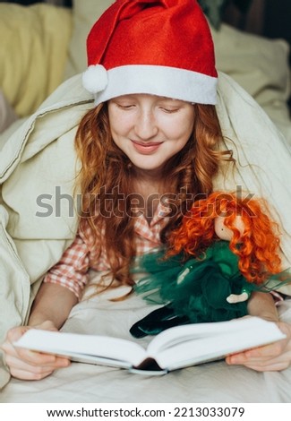 Red-haired girl in santa claus hat reading a book to her doll