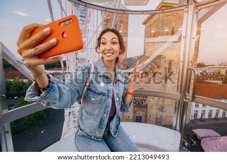 A traveler girl takes a selfie photo for social networks at an amusement fair inside a Ferris wheel booth on the old European city square Royalty-Free Stock Photo #2213049493