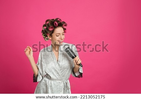 Beautiful young woman in silk bathrobe with hair curlers singing into hairbrush on pink background. Space for text