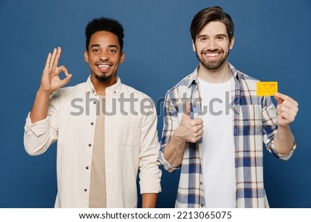 Young two friends men wear white casual shirts together looking camera hold in hand mock up of credit bank card show ok thumb up isolated plain dark royal navy blue background People lifestyle concept