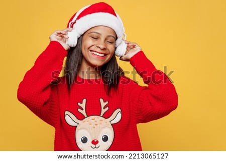 Merry happy smiling little kid teen girl 13-14 year old wear red xmas sweater with deer Santa hat headphones posing listen music isolated on plain yellow background Happy New Year 2023 holiday concept