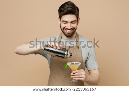 Young happy man barista barman employee in brown apron work in bar pub club pours alcohol make cocktail in martini glass isolated on plain pastel light beige background Small business startup concept Royalty-Free Stock Photo #2213065167