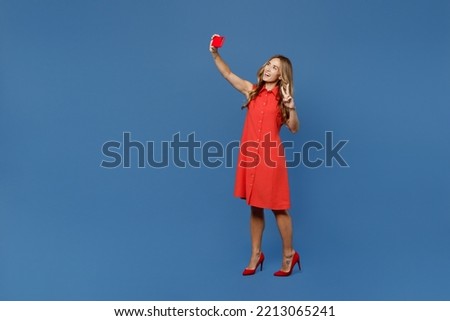 Full body young woman 30s she wear red dress doing selfie shot on mobile cell phone post photo on social network show v-sign isolated on plain dark royal navy blue background. People lifestyle concept