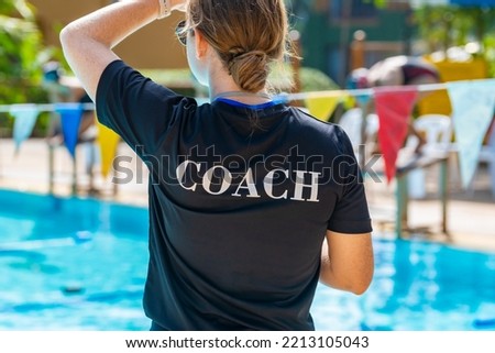 Back view of female swimming coaches, wearing COACH shirt, working at an outdoor swimming pool Royalty-Free Stock Photo #2213105043