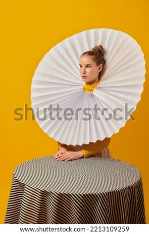 Dinner party. Young indifferent girl in giant jabot collar or neckwear and yellow tights isolated over yellow background. Contemporary art, weird beauty, avant-garde fashion. Royalty-Free Stock Photo #2213109259